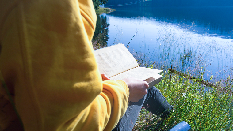 Person sitter i gräset precis vid vattnet och läser en bok. 